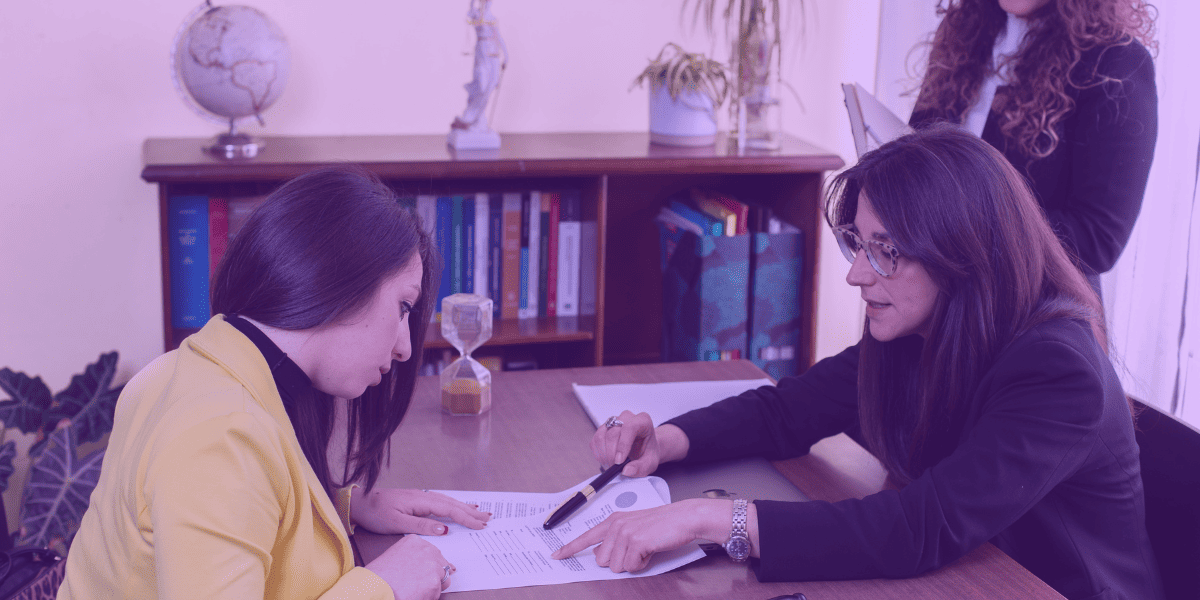 quem tem direito a gratuidade de justiça A imagem mostra duas mulheres em uma conversa séria em um ambiente de escritório. Uma delas, vestindo uma jaqueta amarela, está sentada à mesa, analisando documentos, enquanto a outra, com um olhar focado, a orienta e aponta informações em um papel. Ao fundo, uma terceira mulher observa, segurando uma pasta, o que sugere que está acompanhando a reunião. Esse cenário ilustra uma consulta legal, onde a questão da gratuidade de justiça pode ser discutida.