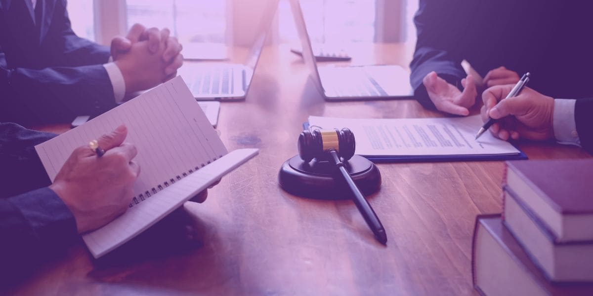 Lei de Falência A imagem retrata uma mesa de reunião com documentos jurídicos, notebook e um martelo judicial no centro, enquanto mãos de profissionais, em trajes formais, fazem anotações e discutem questões legais sobre a Lei de Falência e Recuperação Judicial.